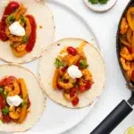 "Plate of shrimp fajitas with colorful bell peppers, onions, and lime wedges, served on a tortilla and topped with sour cream, alongside a cast-iron skillet with more fajita filling."